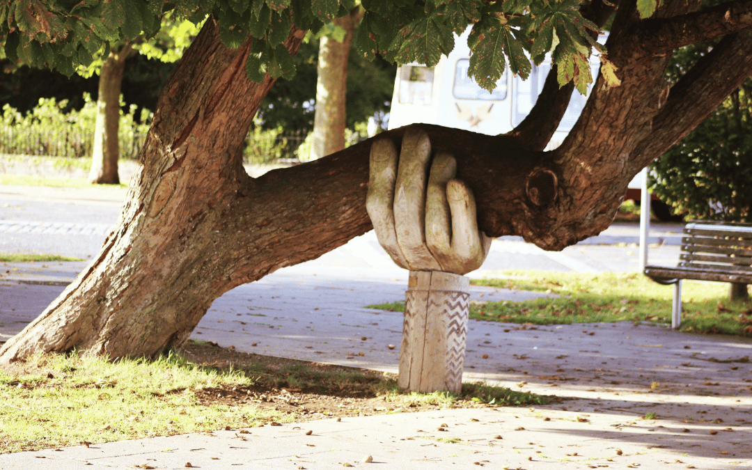 wooden sculpture of hand supporting horizontal branch of leaning tree with road and caravan in background.